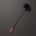 A long-handled grooming brush with a dark bristle head and a textured reddish-brown handle, photographed diagonally on a dark gray background.