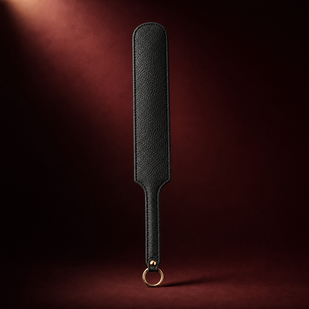 Black pebbled leather impact paddle with a gold-tone hanging ring on a deep burgundy background.